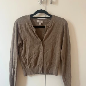 Beige kofta från H&M - Mörkbeige kofta från H&M med rund halsringning och knappar framtill. Perfekt att slänga över ett linne eller t-shirt. Mjuk och tunn, passar året runt.