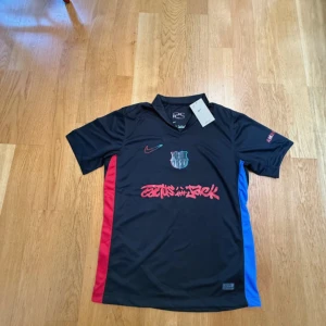 FC Barcelona x Cactus Jack Nike tröja - Snygg svart FC Barcelona x Cactus Jack fotbollströja från Nike med röda och blå sidopaneler, klubbmärke på bröstet och Cactus Jack-tryck i rött framtill. Tillverkad i lätt och ventilerande polyester med korta ärmar och V-ringning.