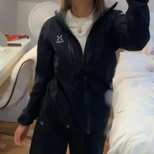 Svart vindjacka från Haglöfs - Säljer en svart vindjacka från Haglöfs med huva och dragkedja framtill. Jackan har en bröstficka med dragkedja och reflekterande detalj på ärmen. Perfekt för blåsiga dagar och outdoor-aktiviteter. Snygg och funktionell design.