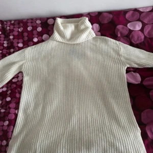 Vit stickad polotröja med ribbad struktur - Säljer en vit stickad polotröja med ribbad struktur och lång ärm. Tröjan har en hög, mysig polokrage och är perfekt för kyliga dagar. Enkel och stilren design som är lätt att matcha med olika outfits.