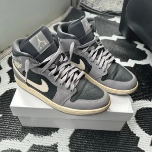 Nike Air Jordan 1 Mid grå/svart - Snygga Nike Air Jordan 1 Mid sneakers i grått och svart skinn med beige swoosh och sula. Klassisk mid-top modell med snörning och ikoniska Air Jordan-loggan på sidan. Perfekta för dig som vill ha en stilren och tidlös look.