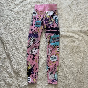 Färgglada leggings från Stronger XS - Supercoola leggings från Stronger i storlek S med serietidningsinspirerat mönster i rosa, blått, gult och svart. Hög midja och stretchigt material i polyester och elastan. Perfekta för dig som vill sticka ut på gymmet eller i vardagen!