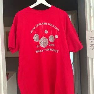 Röd New England College t-shirt - Röd t-shirt med tryck från New England College Holiday Tournament 2018. Framsidan har ett coolt basketbollmotiv i vitt och text. Klassisk passform och rund hals, perfekt för dig som gillar sportig stil.