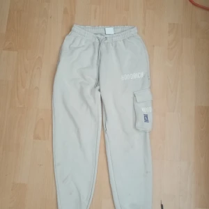 Beige mjukisbyxor från Hoodrich XS - Beiga mjukisbyxor från Hoodrich med elastisk midja och snörning. Byxorna har en stor cargoficka på benet med Hoodrich-logga och tryck, samt relaxed fit. Perfekta för chill dagar eller streetwear-stil., modellen på dessa hoodrichbyxor säljs inte längre