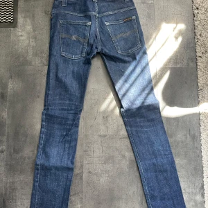 Nudie Jeans blå jeans straight fit - Snygga blå jeans från Nudie Jeans i klassisk straight fit. Tillverkade i 100% ekologisk bomull med coola kontrastsömmar på bakfickorna och läderpatch i midjan. Perfekta för dig som gillar stilrena och hållbara jeans.