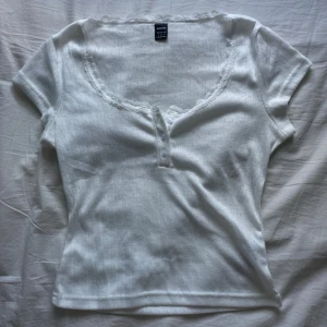 Brandy Melville inspirerad topp - Supersöt vit topp från SHEIN med spetskant och små knappar framtill. Tunt, lätt och skönt ribbat tyg med korta ärmar. Helt oanvänd!