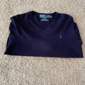 Mörkblå v-ringad tröja Polo Ralph Lauren - Klassisk mörkblå v-ringad tröja från Polo Ralph Lauren med broderad grön logga på bröstet. Tröjan är långärmad och har ribbade muddar vid ärmslut och nederkant. Perfekt för dig som gillar stilren och tidlös design.