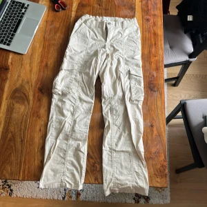 Beiga cargobyxor från Lindex, stl 152 - Beiga cargobyxor från Lindex med elastisk midja och flera praktiska fickor på sidorna. Byxorna är raka i modellen och tillverkade i mjuk bomull. Något slitna i benslut, annars toppenskick