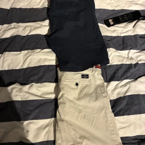 Två par klassiska shorts herr - Säljer två par stilrena shorts från Dressmann. Ett par är marinblå med randig midja, det andra är grå  med röd och blå detalj i midjan. Båda har klassisk passform, fickor och är perfekta för sommaren. Sköna och snygga till t-shirt eller skjorta.