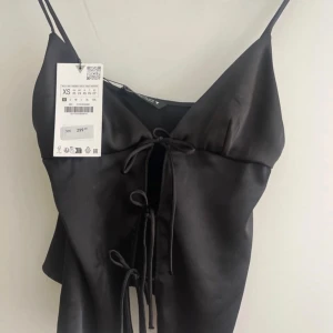 Svart satinlinnetopp med snörning - Supersnygg svart linne-topp i satin från zara med tunna axelband och trendig snörning framtill. Perfekt för att lyfta vilken outfit som helst med sin glansiga finish och coola detaljer. Prislappen sitter inte längre kvar. Använd en gång