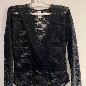 Svart spetsig omlott bodysuit KappAhl - Svart långärmad bodysuit från KappAhl i transparent spets med blommigt mönster. Djup v-ringning med omlott och fina spetsdetaljer vid ärmslut. Perfekt för att skapa en edgy och snygg look.