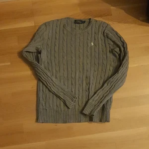 Grå kabelstickad tröja Polo Ralph Lauren - Kabelstickad grå tröja från Polo Ralph Lauren med klassisk rund hals och broderad logga på bröstet. Långärmad modell i mjuk bomull, perfekt för lager-på-lager och chill stil. Snygga ribbade muddar vid ärmslut och nederkant.