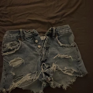 Rippade jeansshorts från ZARA - Säljer ett par blå, högmidjade jeansshorts från ZARA med rejält slitna detaljer och fransig kant. Shortsen har flera stora hål och råa kanter för en riktigt edgy look. Perfekta för dig som gillar streetstyle och vill sticka ut i sommar.