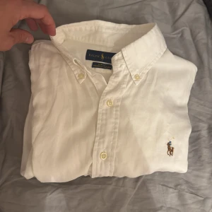 Vit skjorta från Ralph Lauren - Klassisk vit skjorta från Ralph Lauren med broderad logga på bröstet. Skjortan har button-down krage och knäppning framtill. Tillverkad i bomull och har en stilren, tidlös look som passar till många olika outfits. Storlek S