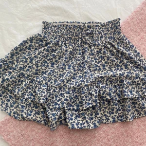Blommig volangkjol med smock - Supersöt kort kjol med blått blommönster på vit botten. Kjolen har smock i midjan med knytband och flera volanger som ger en flowy känsla. Perfekt för dig som gillar romantisk och feminin stil.