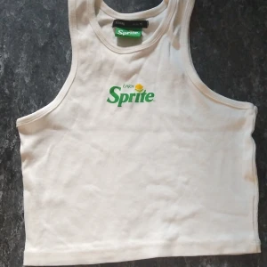 Vit croppad Sprite linne från Sinsay - Säljer en vit croppad linne-topp från Sinsay med Sprite-logga tryckt framtill i grönt och gult. Toppen är ärmlös med klassisk linneform och tillverkad i mjuk bomull. Perfekt för dig som gillar coola loggor och vill sticka ut lite extra.