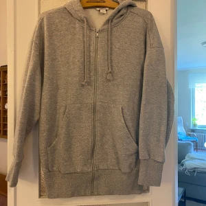 Grå hoodie med dragkedja från Monki - Mysig grå hoodie från Monki i storlek M. Tröjan har huva med snörning, dragkedja framtill och två stora fickor. Tillverkad i mjuk bomullsblandning och har en relaxed passform med långa ärmar. Perfekt för chill dagar eller när du vill vara extra bekväm. Storlek M men skulle säga att den passar L också.