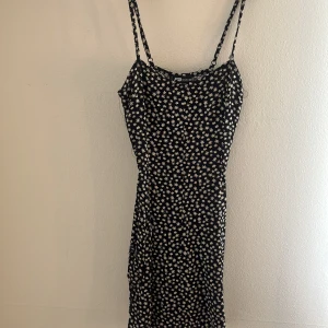 Svart blommig kortklänning FB Sister - Supersöt svart kortklänning från FB Sister med vita och gula blommor, smala axelband och snörning framtill. Perfekt för varma dagar och ger en riktigt somrig vibe. Sitter figurnära och har en feminin siluett.