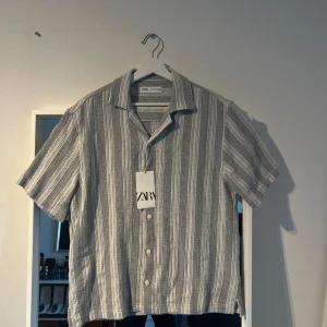 Randig kortärmad skjorta från Zara - Snygg kortärmad skjorta från Zara i grått och vitt med vertikala ränder. Aldrig andvänd med tagen fortfarande kvar!