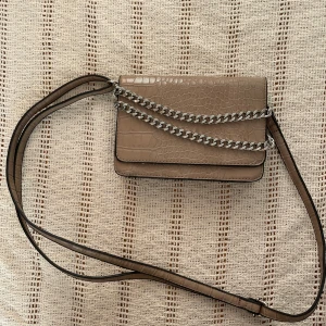 Beige axelväska med kedja från Don Donna - Snygg beige axelväska från Don Donna med croco-mönstrad yta och silverfärgad kedjedetalj. Insidan har coolt prickigt foder och flera fack, inklusive ett med dragkedja. Perfekt storlek för mobilen och det viktigaste.