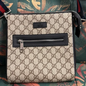 Gucci axelväska med GG-mönster - Snygg axelväska från Gucci i beige och brunt GG-mönster. Väskan har svart läderdetalj runt dragkedjan framtill och en bred, justerbar axelrem i rött och marinblått. Tillverkad i canvas och skinn, med innerfack och dragkedja för extra säkerhet.