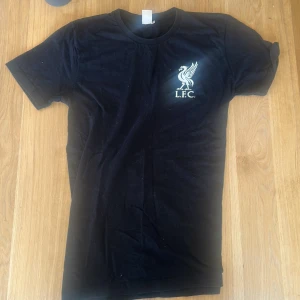 Svart Liverpool t-shirt med tryck - Svart t-shirt från Liverpool med klassiskt vitt tryck. T-shirten har rund halsringning och korta ärmar. Perfekt för dig som vill visa ditt stöd för Liverpool FC på ett stilrent sätt.