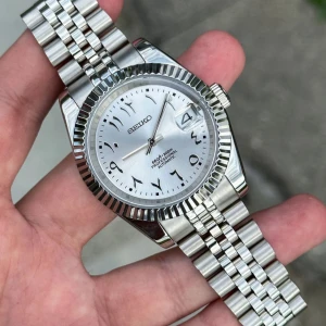 Seiko mod datejust Arabic silver - Snygg Seiko automatisk klocka i silver med klassisk jubilee-länk och räfflad bezel. Urtavlan är silverfärgad med arabiska siffror och datumfönster vid klockan tre. Robust design i rostfritt stål, perfekt för dig som gillar stilrena accessoarer.
