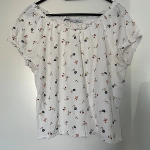 Blommig topp, Lager 157, L - Vit topp med blommigt mönster och korta ärmar från Lager 157. Blusen har en rynkad halsringning och en lätt textur. 100% bomull. Nyskick.  Mått: Längd ca 50 cm Byst ca 52 cm Midja ca 48 cm
