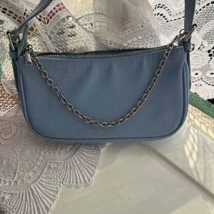 Blå axelväska med kedjedetalj - Snygg ljusblå axelväska i syntet med croco-mönster och silverfärgad kedja framtill. Väskan har dragkedja upptill och en stilren, avlång form som passar perfekt till din outfit. Enkel att bära över axeln och ger en trendig touch.