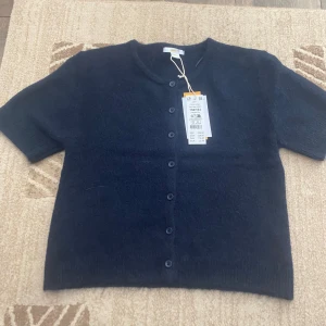 Mörkblå stickad kofta från Gina Tricot - Kortärmad, mörkblå stickad kofta från Gina Tricot med rund halsringning och knappar framtill. Koftan har en mjuk och fluffig känsla, perfekt för lager-på-lager och en chill look. Enkel att matcha med både jeans och kjol.