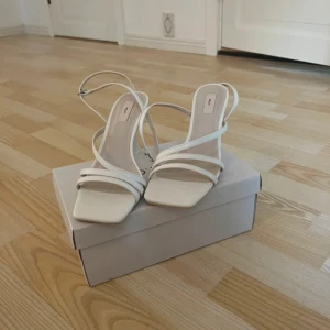 Vita sandaler med smala remmar och klack - Stilrena vita sandaler med smala remmar och fyrkantig tå. Skorna har en elegant hög klack och justerbar rem runt ankeln. Perfekta för dig som gillar minimalistisk och trendig design. Tillverkade i syntetmaterial. Ny köpta aldrig använda 