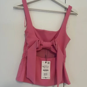 Rosa linnetopp med stor rosett XS - Supersöt rosa linnetopp med breda axelband och en stor rosett framtill. Toppen har en öppen detalj under rosetten och är figursydd för en snygg passform. Perfekt för dig som vill sticka ut med en färgglad och lekfull look. Det finns smink på framsidan av tröjan som definitivt kommer att gå bort i tvätten!