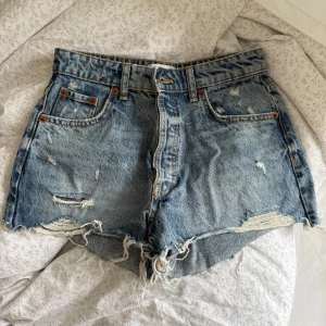 Blå jeansshorts från Zara - Säljer ett par blå jeansshorts från Zara med råa kanter och slitna detaljer. Klassisk femficksmodell i denim med fransig nederkant och coola slitningar framtill. Perfekta för sommaren och riktigt snygga till sneakers eller sandaler.