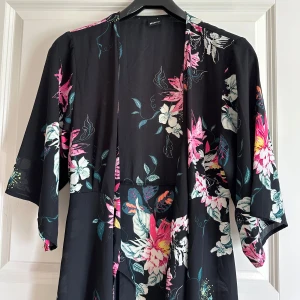 Svart blommig långklänning med knytband Gina Tricot morgonrock - Svart långklänning från Gina Tricot med färgglada blommor i rosa, vitt och grönt. Klänningen har trekvartsärmar och ett knytband i midjan för en snygg siluett. Lätt och flowigt material, perfekt för varma dagar. Kan även användas som en tunn morgonrock.