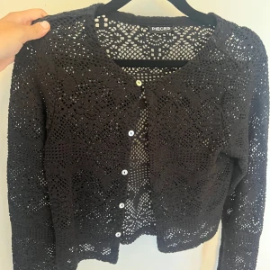Svart virkad kofta från Pieces - Säljer en svart virkad kofta från Pieces med fina hålmönster och vita knappar framtill. Koftan har lång ärm och är perfekt att slänga över en topp för en chill och snygg look. Passar både till jeans och kjol. Aldrig använt.