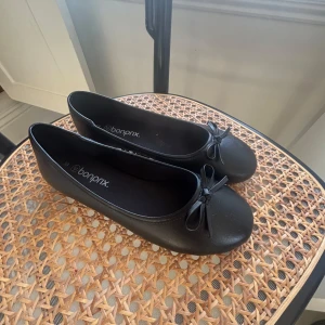 Svarta ballerinaskor från Bonprix - Klassiska svarta ballerinaskor från Bonprix med rund tå och liten rosett framtill. Skorna har platt sula och är tillverkade i syntetmaterial, vilket gör dem lätta och smidiga att bära. Perfekta för en stilren och enkel look.