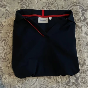 Mörkblå pikétröja Sea Ranch - Snygg mörkblå pikétröja från Sea Ranch i storlek XXL barnstorlek vilket motsvarar S/M. Tröjan har röda och vita detaljer vid krage och ärmslut, klassisk krage och korta ärmar. Perfekt för en stilren och sportig look. Materialet känns som bomull och är mjukt mot huden.