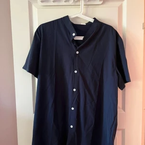 Mörkblå kortärmad henley-skjorta - Snygg mörkblå henley-skjorta med korta ärmar och vita knappar framtill. Skjortan har en enkel och stilren design med ståkrage och är tillverkad i ett lätt bomullstyg som passar perfekt till varma dagar.