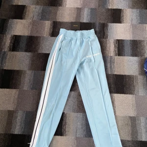 Palm Angels ljusblå träningsbyxor - Palm Angels ljusblå träningsbyxor med vita och svarta ränder längs sidorna. Byxorna har elastisk midja och raka ben, samt logga tryckt på vänster sida. Perfekta för en sportig och trendig look.