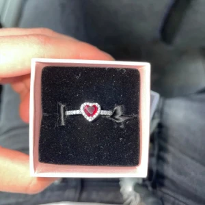 Pandora silverring med röd hjärtsten - Säljer en snygg silverring från Pandora med en röd hjärtformad sten i mitten, omgiven av små glittrande stenar. Ringen har en elegant och romantisk vibe, perfekt för dig som gillar detaljer och vill sticka ut lite extra.