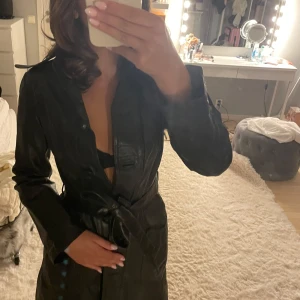 Svart skinn trenchcoat med bälte - Säljer en svart trenchcoat i skinn med snyggt bälte i midjan. Jackan har klassisk krage, knappar framtill och långa ärmar. Perfekt för dig som vill ha en cool och tidlös look. Passar lika bra till jeans som till klänning. Köpt för ca 800kr, aldrig använd! 