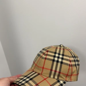Burberry rutig keps i beige och rött - Snygg Burberry keps med klassiskt rutigt mönster i beige, svart och rött. Kepsen har böjd skärm och justerbar rem bak. Tillverkad i bomull med svart insida. Perfekt för dig som vill ha en ikonisk accessoar med lyxig känsla.