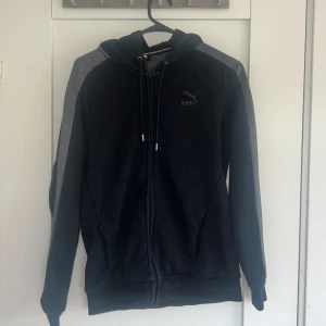 Svart hoodie från Puma med dragkedja - Svart hoodie från Puma med silver paneler längs ärmarna och diskret Puma-logga på bröstet. Tröjan har huva med snörning, hel dragkedja och två fickor framtill. Materialet är mjuk bomullsmix, perfekt för chill eller träning.