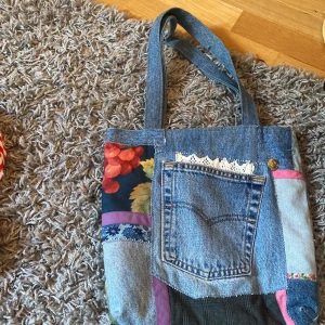Patchwork shoppingväska i jeans - Unik shoppingväska i patchwork-stil med jeansdetaljer, ficka framtill och blommiga tyginslag i grönt, blått och rött. Perfekt för dig som gillar färg och personliga accessoarer. Nypris:vet ej