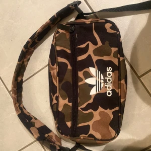 Camouflage axelväska från Adidas - Säljer en snygg axelväska från Adidas med klassiskt camouflagemönster i brunt, grönt och svart. Väskan har ett stort fack med dragkedja och justerbar axelrem. Adidas-logga i vitt på framsidan ger en sportig vibe. Perfekt för att bära med sig det viktigaste.