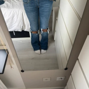 Blå bootcut jeans med hål - Säljer ett par blå jeans med bootcut passform och stora hål vid knäna. Jeansen har klassisk femficksdesign och är tillverkade i jeansmaterial. Perfekt för dig som gillar en avslappnad och trendig stil.