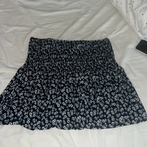 Blommig kortkjol från Gina Tricot - Supersöt kortkjol från Gina Tricot med mörkblå botten och vita blommor. Kjolen har en smockad midja som ger en skön och flexibel passform. Materialet är mjukt och lätt, perfekt för varma dagar. Passar dig som gillar en feminin och chill stil.