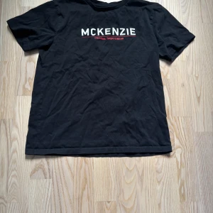 Svart McKenzie t-shirt med tryck - Svart t-shirt från McKenzie med vit och röd logga tryckt på bröstet. Klassisk rund hals och korta ärmar. Tillverkad i mjuk bomull som är skön mot huden. Perfekt för dig som gillar enkel och stilren streetwear.