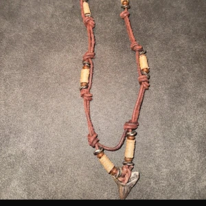 Halsband med hajtand och bruna detaljer - Unikt halsband med brun mockaliknande snodd, dekorerat med beige och bruna pärlor samt en cool hajtandsberlock i grått. Knytningar längs bandet ger en avslappnad vibe. Perfekt accessoar för dig som gillar en boho eller surf-inspirerad stil.