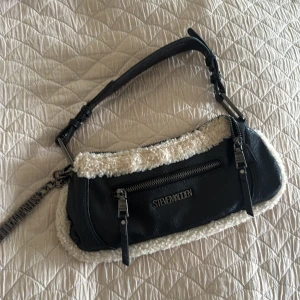 Svart axelväska med teddy från Steve Madden - Cool svart axelväska från Steve Madden med mjuka teddy-detaljer i beige längs kanterna. Väskan har silverfärgade metalldetaljer, justerbar axelrem och dragkedja både fram och upptill. Insidan har extra fack med dragkedja. Perfekt storlek för det viktigaste.